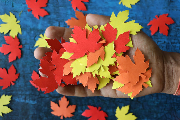 照片中，一个认不出来的人拿着一把自制的秋叶剪影剪影，红色、橙色和黄色的枫叶形状的剪影，盖在皱巴巴的蓝色背景上图片下载