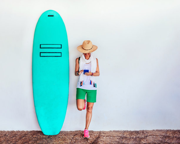 一个女人靠在白色的墙上，旁边是一块绿色的冲浪板。她拿着一部手机。图片下载