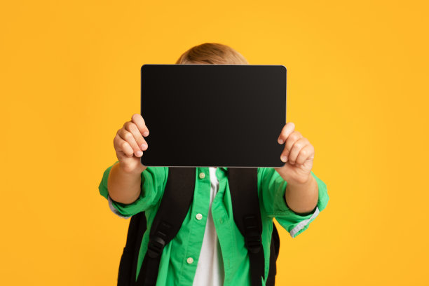 无法辨认的金发少年小学生显示平板电脑与空屏幕，孤立在橙色背景图片下载