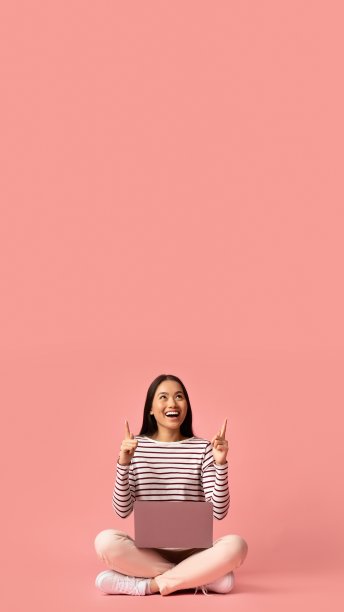 检查这个。兴奋的亚洲女性坐在笔记本电脑和指向上方的复制空间，在粉红色的工作室背景，开朗的年轻韩国女性显示你的文本或设计的自由空间图片下载