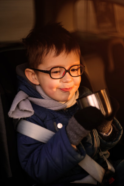 3-4岁的小男孩，戴着眼镜，坐在一辆车里，在夕阳的橙色光线下微笑着用保温杯喝水图片下载