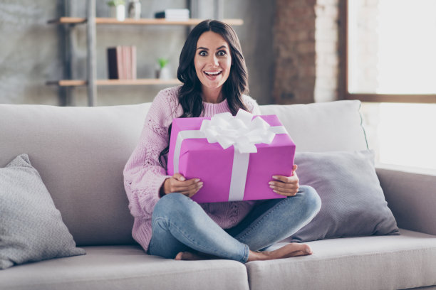 惊喜快乐的女人的全身照片，坐在沙发里，在屋里，在家里，抱着大礼盒图片下载