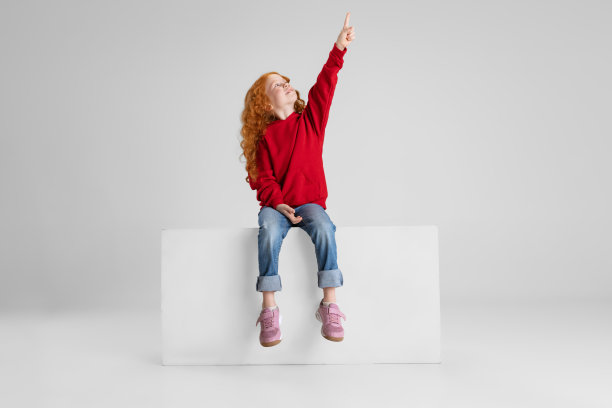 小红发卷发女孩的肖像，孩子指着某个地方孤立的灰色工作室背景。图片下载