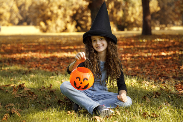 可爱的微笑女孩，戴着女巫帽，坐在草地上，手里拿着一个橙色的大锅，看着相机。图片下载