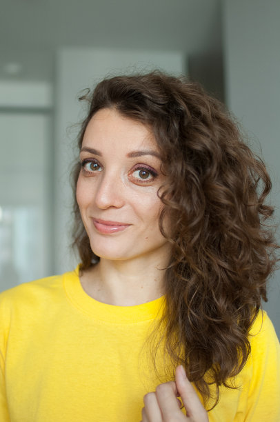 美丽的卷发女孩穿着黄色t恤在她的公寓里看着摄像机，微笑着摆姿势，快乐的人们概念图片下载