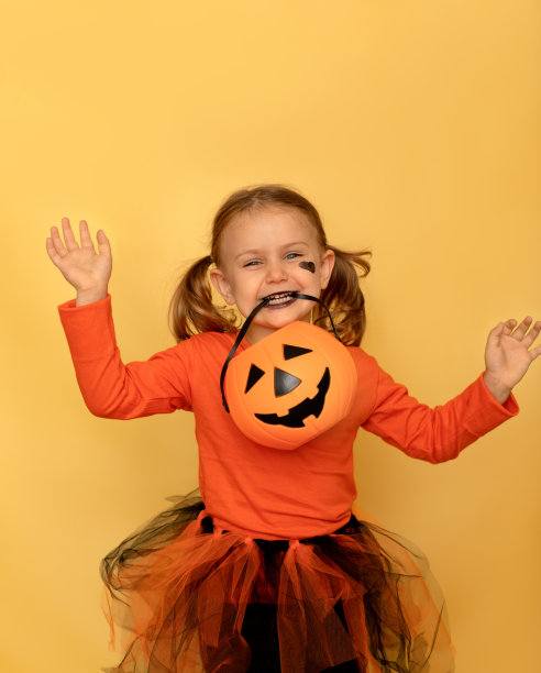 有趣的万圣节派对。工作室拍摄黄色孤立背景。微笑强烈情感的孩子在万圣节橙色女巫服装，化妆黑色格林和南瓜。万圣节,狂欢节。空间的文本。垂直图片下载