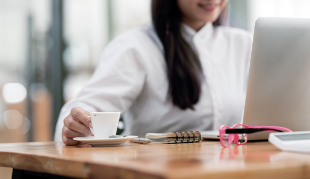 一名女子在办公室用笔记本电脑工作时，手握一杯咖啡。图片下载