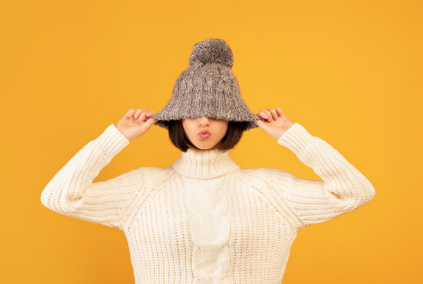 漂亮的年轻女子拉下羊毛帽，露出亲吻的嘴唇，在黄色背景下对着镜头佯装和摆姿势图片下载