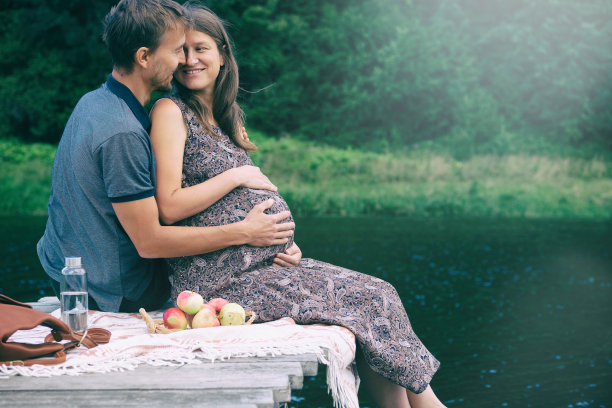 一个孕妇和她的丈夫一起坐在湖边的木码头上。一个年轻家庭即将怀孕的偷拍照片。幸福的情侣在夏日户外野餐。图片下载