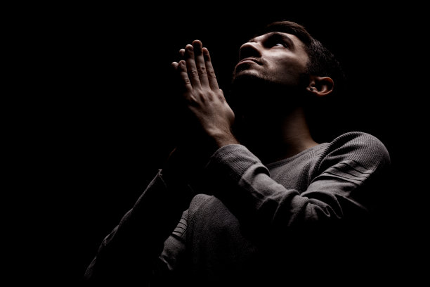 一个严肃自信的年轻人在黑暗的房间里祈祷。从上面照亮，向上看图片下载
