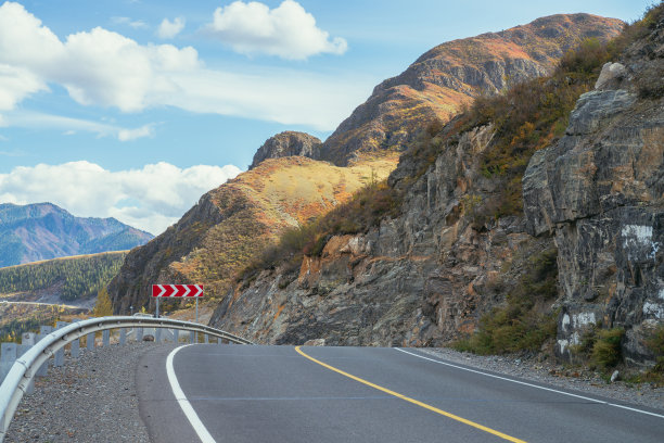 阳光下的山石公路，秋色斑斓。明亮的高山风景与秋天色彩的山路。公路在山区的秋天时间。路沿着美丽的石壁。图片下载