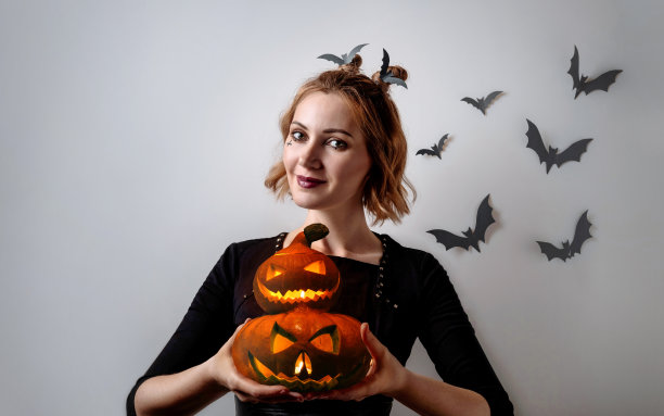 身着万圣节妆容的金发女吸血鬼手中拿着灰色背景上雕刻的南瓜。红嘴唇和烟熏妆的时尚女孩。图片下载