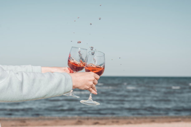 两杯玫瑰酒在女人的手中，在天空和海岸的衬托下。图片下载