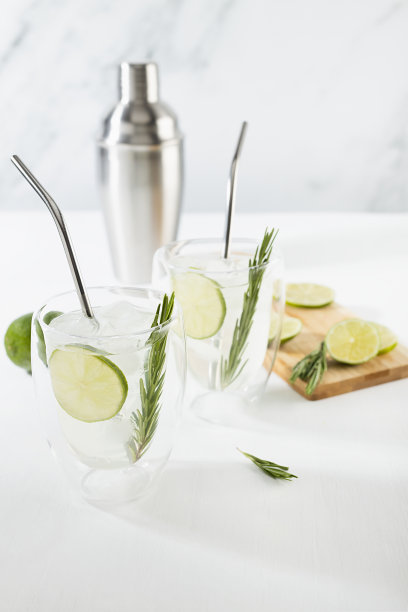 夏日清凉的莫吉托鸡尾酒搭配冰，酸橙片，绿色迷迭香树枝在透明的杯子和银摇壶，吸管在优雅的阳光白色内部木制桌子，垂直。图片下载