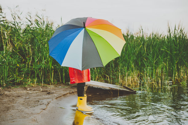 男孩穿着红色雨衣和黄色胶靴站在河边，手里拿着彩虹伞。一名学生静静地站在秋天的湖边。海滨穿着防水衣服的孩子图片下载