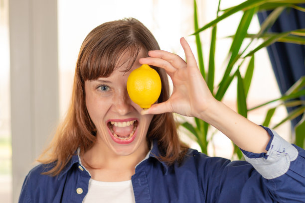 一个年轻漂亮的女人穿着衬衫，手里拿着一个明亮的柠檬，在一扇明亮的窗户和一株绿色的室内植物的叶子的背景下闲逛。有选择性的重点图片下载