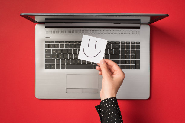 第一个人俯视图的照片，手握白色贴纸纸条与画的微笑的脸在开放的灰色笔记本电脑孤立的红色背景图片下载