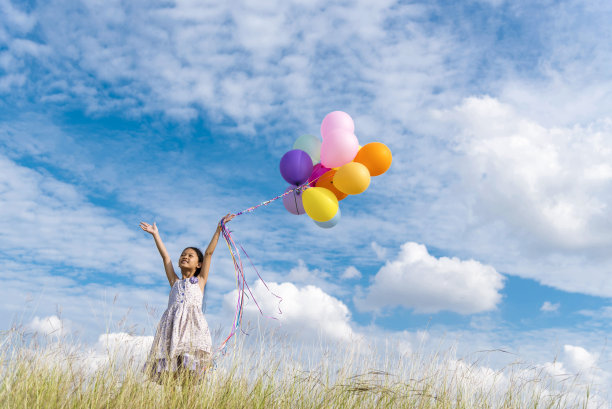 欢快可爱的女孩举着气球在绿色的草地、白云、蓝天上快乐地奔跑。手握着充满活力的气球在生日派对上玩耍快乐的时光，夏天在户外的阳光下图片下载