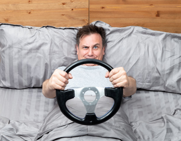 滑稽的人躺在床上，抬起头，拿着前面的汽车方向盘。驾驶者概念随时随地。图片下载