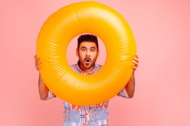 惊讶的络腮胡男人，穿着蓝色休闲衫，拿着游泳圈，惊讶地看着镜头图片下载