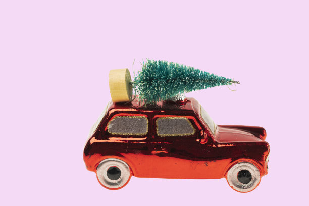 近距离观察玻璃玩具装饰形式的红色汽车人物运送圣诞树孤立在粉红色的背景。图片下载