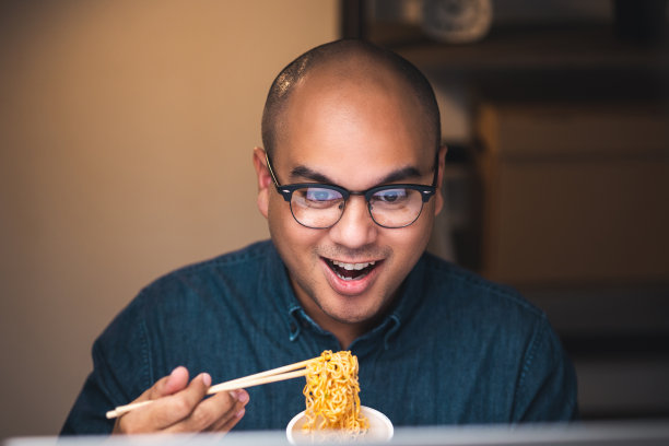 年轻的亚洲自由职业者工作到深夜。晚上他很饿，吃着热腾腾的方便面，在黑暗的办公室里用电脑工作。图片下载