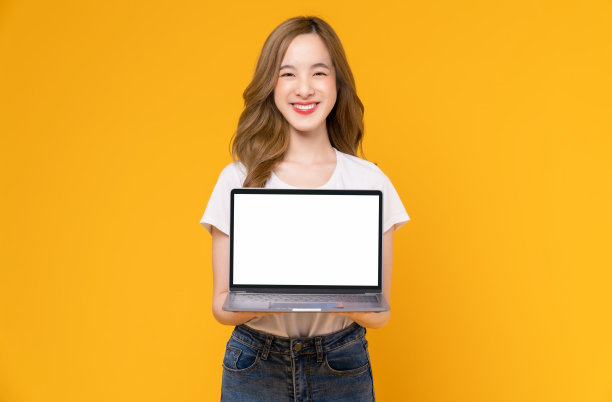 摄影棚拍摄的开朗美丽的亚洲女人在白色t恤和手持笔记本电脑模型的空白屏幕上的黄色背景。图片下载