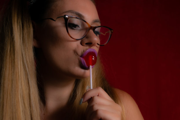 特写的年轻和美丽的女人享受棒棒糖，好看可爱迷人，欢快的马尾辫，微笑，在一个红色的背景。股票摄影。图片下载