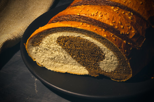 不寻常的两色新鲜面包在陶瓷盘和编织餐巾的黑色背景，照片在强光下低调，特写。图片下载