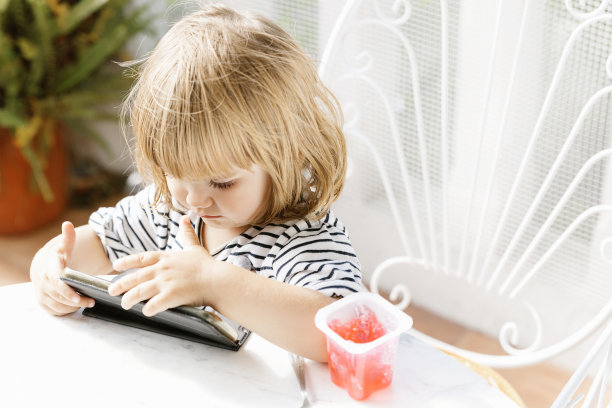 夏天，可爱的小女孩坐在后院的桌子旁，一边看卡通片一边享受美味的果冻甜点图片下载