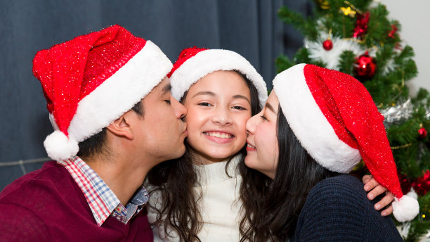 圣诞节那天，爸爸妈妈穿着圣诞老人的毛衣，戴着圣诞老人的帽子，在家里亲吻快乐的女儿。圣诞快乐，节日快乐。圣诞节的庆祝活动图片下载