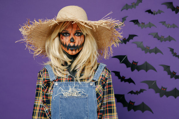 年轻的金发女子带着万圣节化妆面具，头戴草帽，身穿牛仔稻草人装束，在镜头孤立的纯暗紫色背景下写真。庆祝节日派对的概念。图片下载