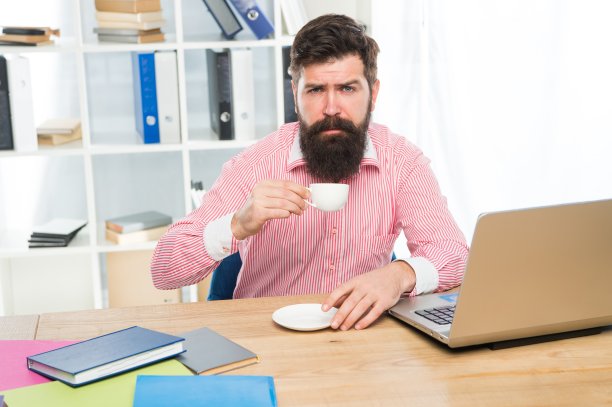 早晨，在现代办公室里，留着时髦胡子的疲惫的文员坐在桌上喝咖啡图片下载
