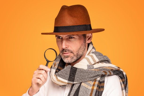 男侦探拿着放大镜，戴着时尚的围巾和棕色的帽子，在黄色的摄影棚背景下摆姿势。图片下载