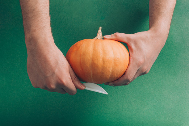创意万圣节创意，男性手拿新鲜南瓜和刀。简约的裁剪和工艺概念，假期的审美可视化。穆迪深绿色背景。图片下载