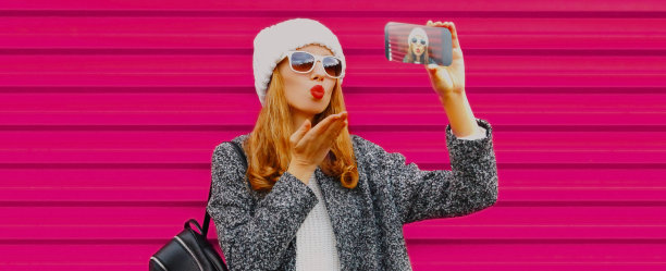 一名年轻女子戴着白色针织帽，以粉色为背景，用智能手机自拍图片下载