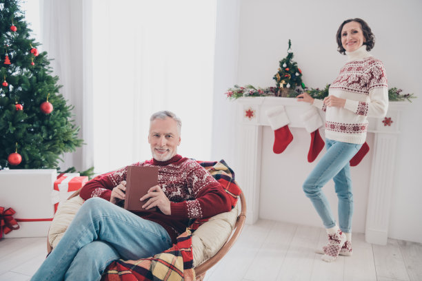 一对甜蜜夫妇的照片，老太太站着，男人看书，穿毛衣，牛仔裤，袜子在家里图片下载