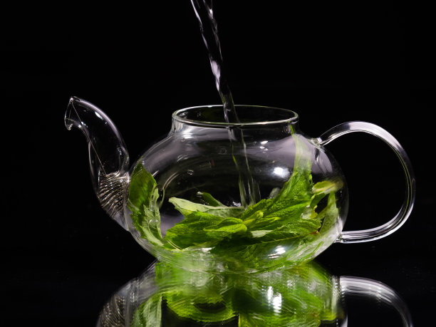 将热水倒入黑色背景的玻璃茶壶中，倒入绿色薄荷茶。草药茶和健康饮料的概念。图片下载
