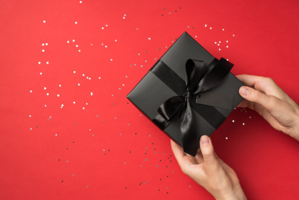 第一人称俯视图照片手握黑色礼品盒与黑色丝带蝴蝶结标签和亮片孤立的红色背景与空白图片下载