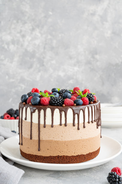在灰色混凝土背景的白色盘子上，放着三块巧克力奶油冻蛋糕，上面点缀着蓝莓、覆盆子和黑莓。有三层不同口味的蛋糕。副本的空间。图片下载