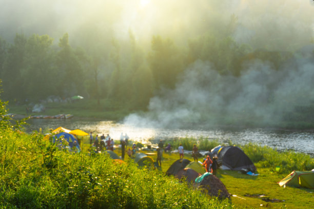 模糊的河边露营。海岸上有帐篷，人们在散步。森林被雾笼罩着。烟从火中蔓延开来。图片下载