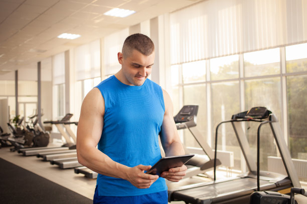 一个年轻英俊的教练在健身房工作的过程中。运动员拿着平板电脑。教练、个人训练、体力锻炼、肌肉发达的身体、快乐的微笑。肖像图片下载