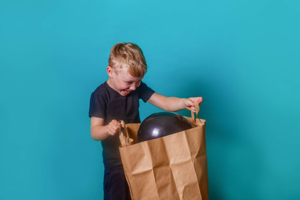 可爱有趣的欧洲小男孩拿着一个沉重的纸袋，蓝色的背景上有黑色的气球。孩子微笑着看着包里的东西。图片下载