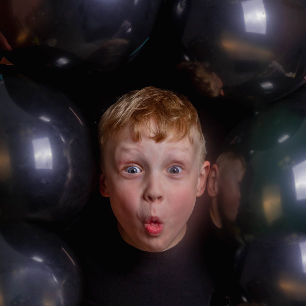 可爱有趣的欧洲小男孩被黑色气球包围着。脸特写。男孩很惊讶，噘起了嘴。背景的气球。图片下载