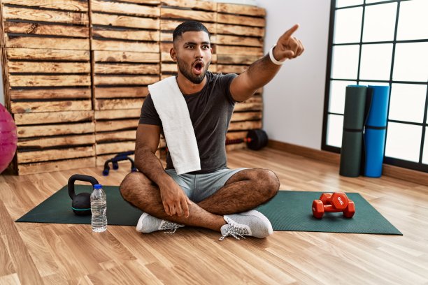 年轻的印度男子坐在训练场的垫子上，手指惊讶地指着前方，张着嘴惊讶的表情，前方有什么东西图片下载