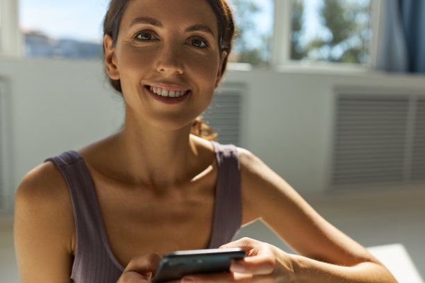 一位30岁的女性，完美的皮肤上有模仿皱纹的自然妆容，对着镜头微笑，在Windows背景下与朋友通过智能手机聊天时被打断图片下载