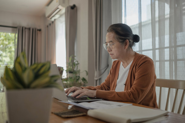 一位亚洲老妇人在家工作，她正在笔记本电脑的键盘上打字。图片下载