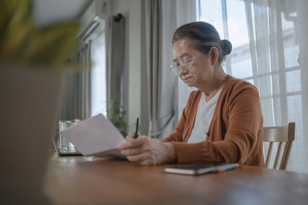 亚洲妇女在餐桌上写便条/信件，回复她另一只手拿着的信件。图片下载