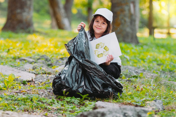 靠近的照片，一个漂亮的年轻女孩坐在公园的地上，拿着一张纸，有一个回收的标志，并把它收进垃圾袋。图片下载