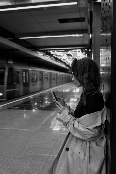身穿米色风衣的女孩博主在地铁站台上用智能手机等车。BW图片下载